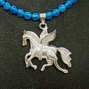 May include: A silver Pegasus pendant necklace with a blue beaded chain. The pendant features a winged horse design, encrusted with small, clear stones. The horse has an orange eye and is set against a black background.