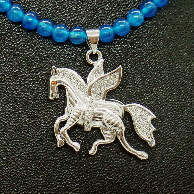 May include: A silver Pegasus pendant necklace with a blue beaded chain. The pendant features a winged horse design, encrusted with small, clear stones. The horse has an orange eye and is set against a black background.
