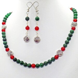May include: A necklace and earring set featuring green and red beads. The necklace has silver-toned Celtic knot accents. The earrings have matching beads and Celtic knot charms. The jewelry is displayed on a white mannequin.