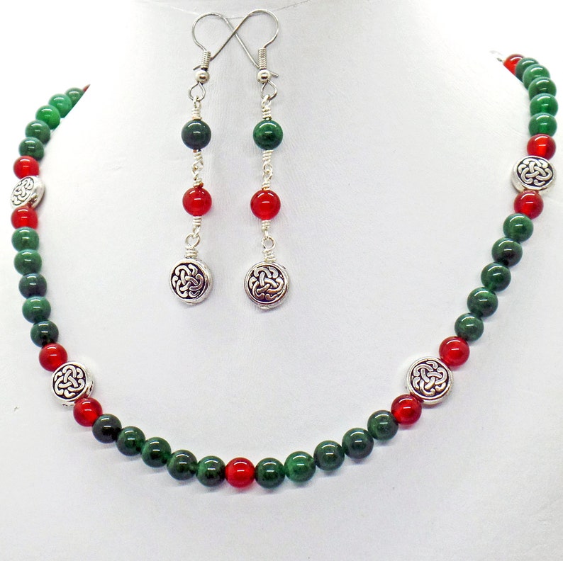 May include: A necklace and earring set featuring green and red beads. The necklace has silver-toned Celtic knot accents. The earrings have matching beads and Celtic knot charms. The jewelry is displayed on a white mannequin.
