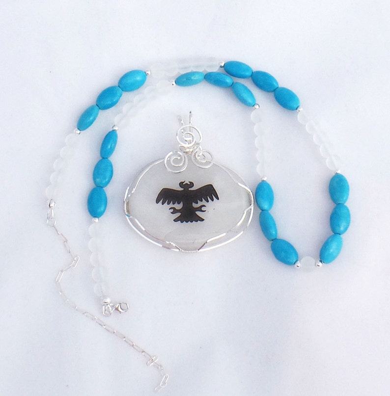 May include: A silver chain necklace with a white oval pendant featuring a black silhouette of a bird. The necklace is strung with turquoise blue beads.