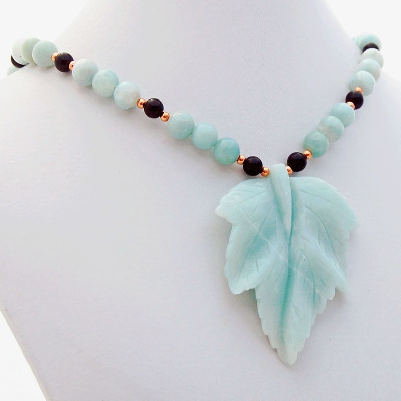 May include: A light blue and black necklace with a carved leaf pendant. The necklace is made of round beads and features a carved leaf pendant made of the same light blue stone.
