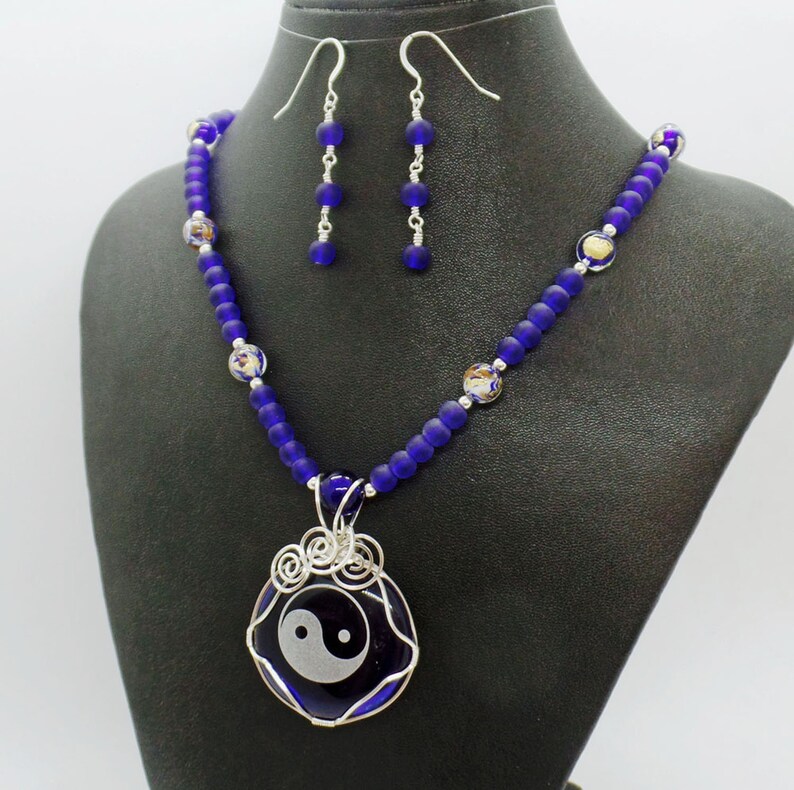 May include: A silver necklace with a blue glass yin yang pendant and matching earrings. The necklace is made with blue glass beads and silver wire.