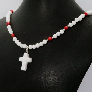 May include: White beaded necklace with a white cross pendant. The necklace features white and red beads, with silver-colored accents. The cross is a simple, classic design. The necklace is displayed on a black mannequin.