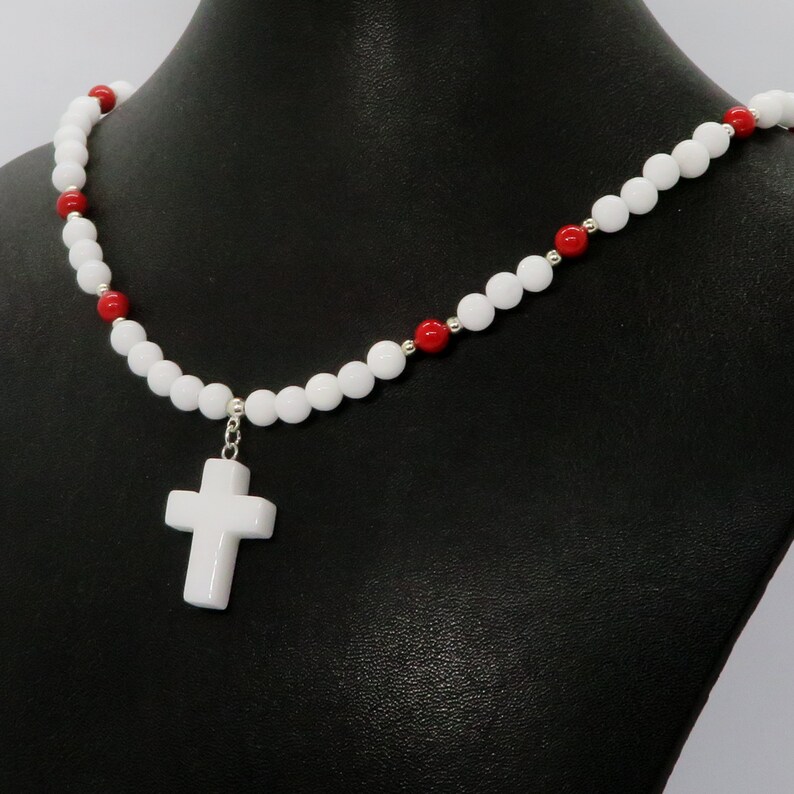 May include: White beaded necklace with a white cross pendant. The necklace features white and red beads, with silver-colored accents. The cross is a simple, classic design. The necklace is displayed on a black mannequin.