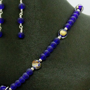 May include: A blue glass beaded necklace with gold flecks and silver accents. The necklace is on a dark green surface.