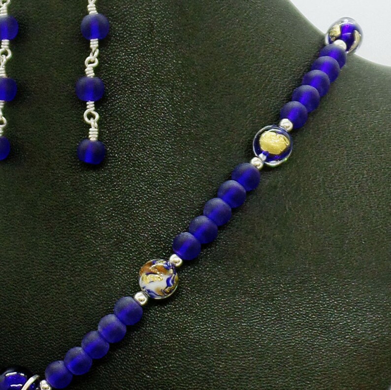 May include: A blue glass beaded necklace with gold flecks and silver accents. The necklace is on a dark green surface.