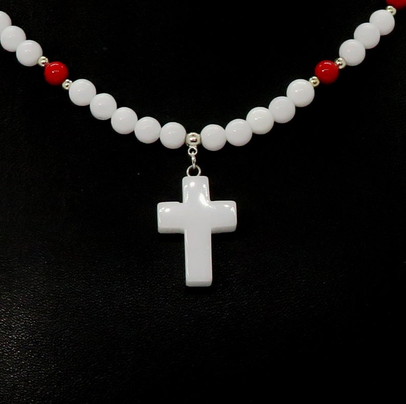 May include: White cross pendant necklace with a beaded design. The necklace features a white cross pendant suspended from a strand of white and red beads. The beads are round and evenly spaced, creating a simple yet elegant design.