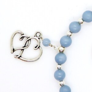 May include: A close-up of a necklace featuring a silver heart-shaped clasp and light blue beads. The beads are interspersed with small silver accents. The necklace is set against a white background.