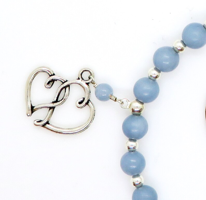 May include: A close-up of a necklace featuring a silver heart-shaped clasp and light blue beads. The beads are interspersed with small silver accents. The necklace is set against a white background.