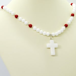 May include: A white beaded necklace with red accent beads and a white cross pendant. The necklace is displayed on a mannequin. The beads are round and the cross is a simple, classic design.