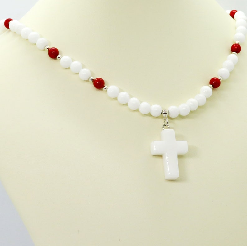 May include: A white beaded necklace with red accent beads and a white cross pendant. The necklace is displayed on a mannequin. The beads are round and the cross is a simple, classic design.