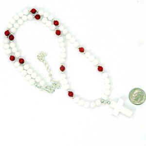 May include: A white beaded necklace with red accent beads and a white cross pendant. The necklace has a silver chain and clasp. A U.S. dime is included for size reference.