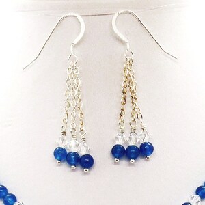 May include: A pair of silver earrings with three strands of delicate chain. Each strand features clear crystals and three deep blue beads. The earrings are displayed against a white background, showcasing the jewelry's elegant design.