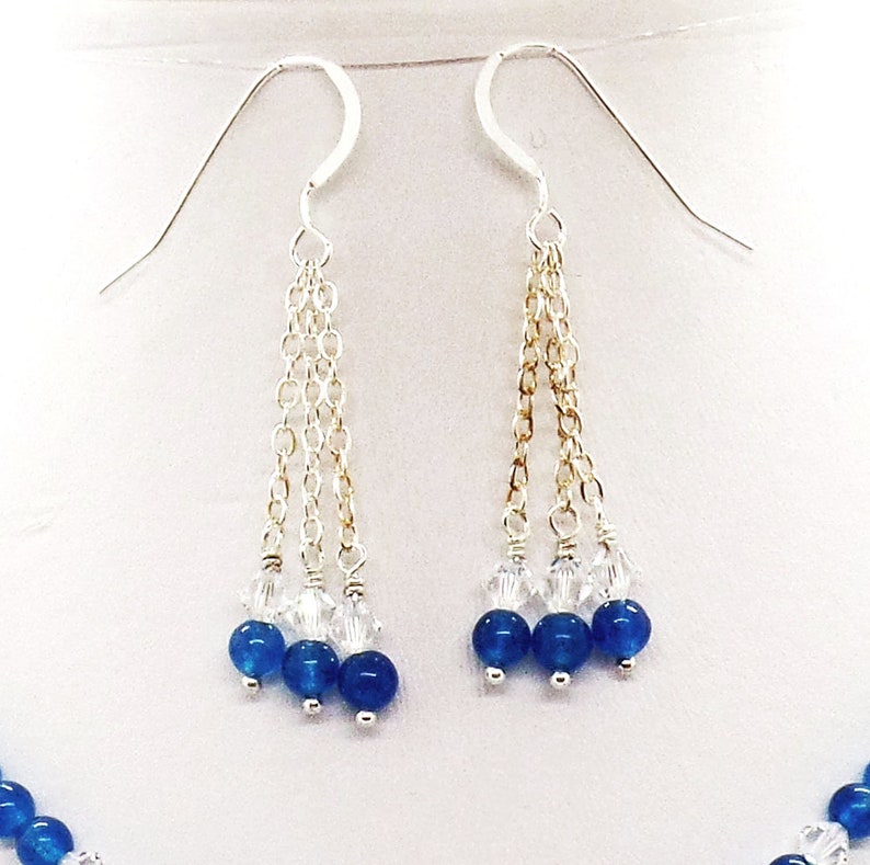May include: A pair of silver earrings with three strands of delicate chain. Each strand features clear crystals and three deep blue beads. The earrings are displayed against a white background, showcasing the jewelry's elegant design.