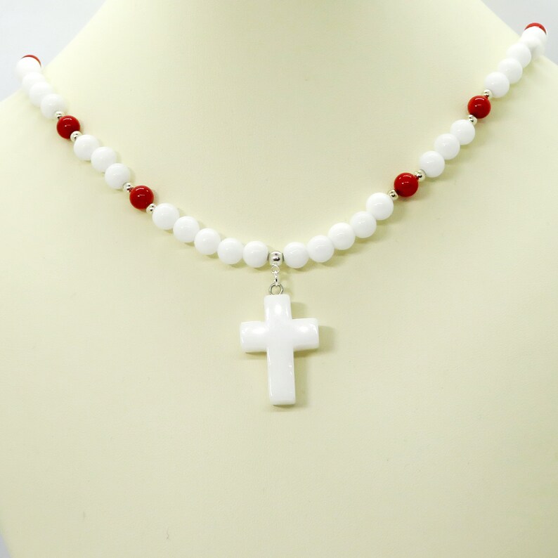 May include: A white beaded necklace with a white cross pendant. The necklace features white and red beads. The cross is a simple, smooth design. The necklace is displayed on a mannequin.