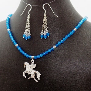 May include: A blue beaded necklace and matching earrings set. The necklace features a silver Pegasus pendant. The earrings have dangling chains with clear and blue beads. The jewelry is displayed on a black mannequin.