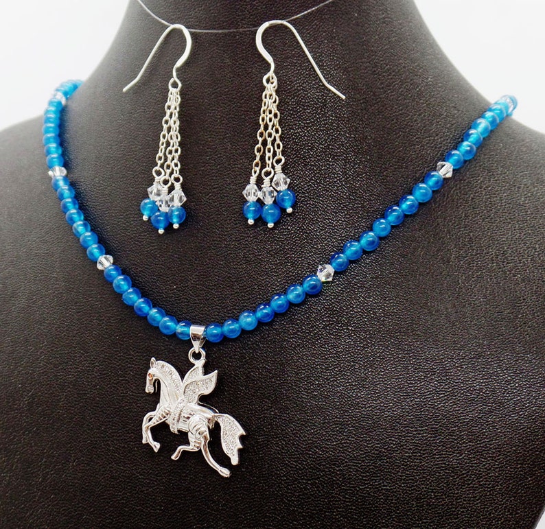 May include: A blue beaded necklace and matching earrings set. The necklace features a silver Pegasus pendant. The earrings have dangling chains with clear and blue beads. The jewelry is displayed on a black mannequin.