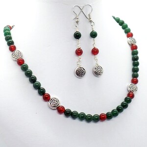 May include: A necklace and earring set featuring green and red beads. The necklace and earrings have silver-colored Celtic knot charms. The earrings hang from silver-colored hooks. The jewelry is displayed on a white background.