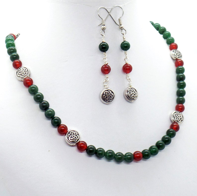 May include: A necklace and earring set featuring green and red beads. The necklace and earrings have silver-colored Celtic knot charms. The earrings hang from silver-colored hooks. The jewelry is displayed on a white background.