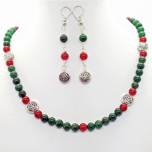 May include: A beaded necklace and earring set featuring green and red beads. The necklace and earrings have silver-toned accents with a Celtic knot design. The earrings dangle from silver hooks.