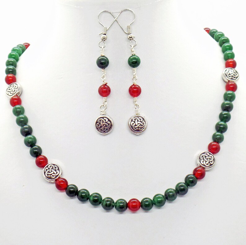 May include: A beaded necklace and earring set featuring green and red beads. The necklace and earrings have silver-toned accents with a Celtic knot design. The earrings dangle from silver hooks.