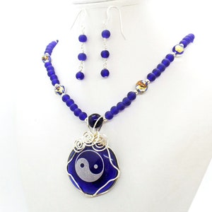 May include: A necklace and earrings set featuring dark blue glass beads and a silver pendant with a yin yang symbol. The pendant is wire wrapped and the earrings are silver with dark blue glass beads.