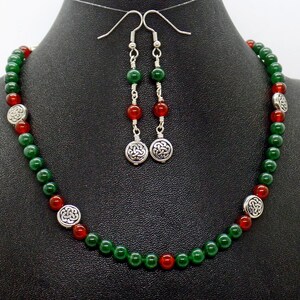 May include: A necklace and earring set featuring green and red beads. The necklace has silver-toned Celtic knot accents. The earrings have matching beads and Celtic knot pendants. The jewelry is displayed on a black mannequin.