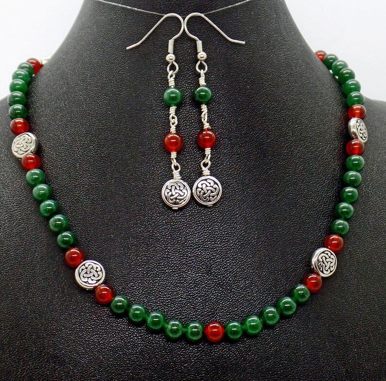 May include: A necklace and earring set featuring green and red beads. The necklace has silver-toned Celtic knot accents. The earrings have matching beads and Celtic knot pendants. The jewelry is displayed on a black mannequin.