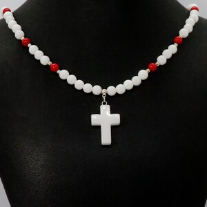 May include: White beaded necklace with a white cross pendant. The necklace features white and red beads, with silver accents. The cross is a simple, classic design. The necklace is displayed against a black background.