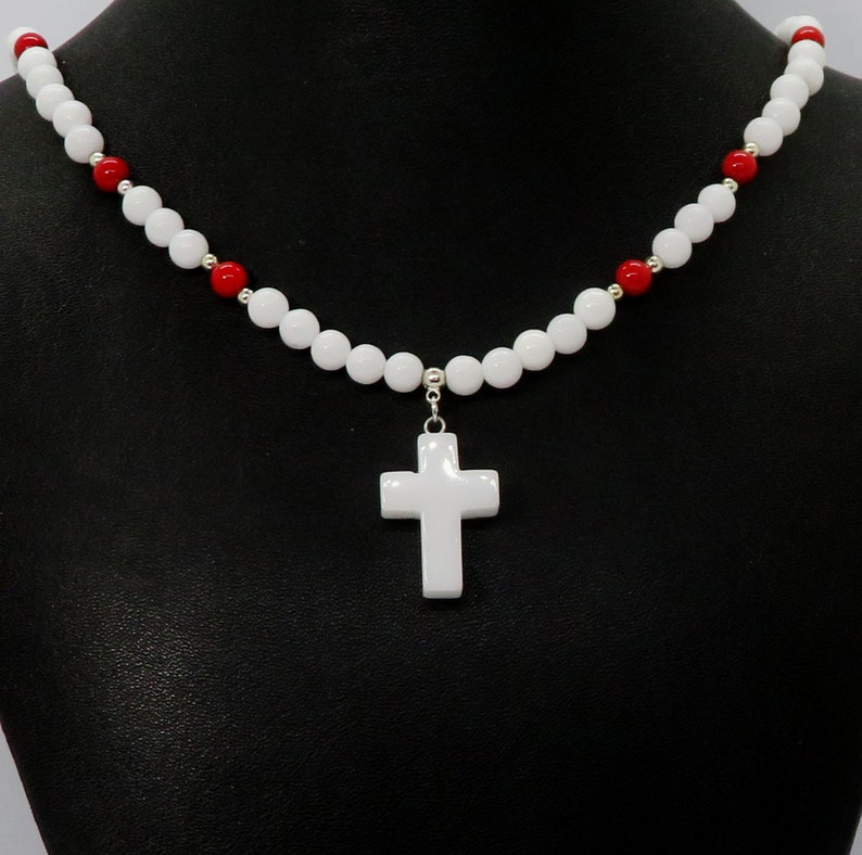 May include: White beaded necklace with a white cross pendant. The necklace features white and red beads, with silver accents. The cross is a simple, classic design. The necklace is displayed against a black background.