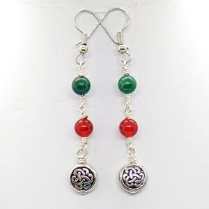 May include: A pair of dangle earrings with green and red beads, connected by silver wire. The earrings have silver Celtic knot charms at the bottom and silver hooks at the top. The beads are round and the charms are circular.