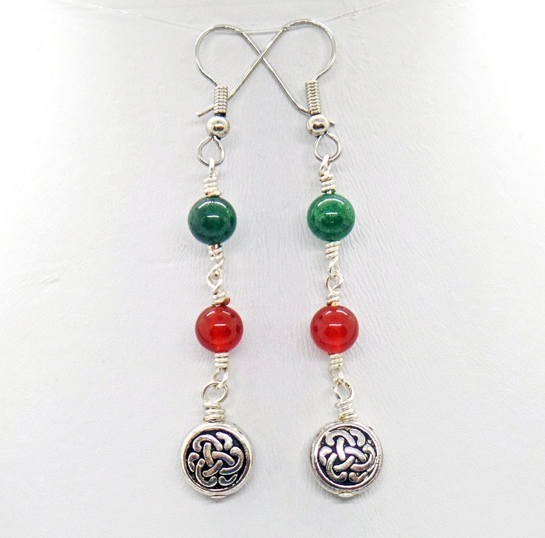 May include: A pair of dangle earrings with green and red beads, connected by silver wire. The earrings have silver Celtic knot charms at the bottom and silver hooks at the top. The beads are round and the charms are circular.