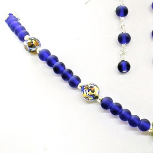 May include: A blue glass bead necklace with silver accents and two matching earrings. The necklace features two larger beads with a white and gold design.