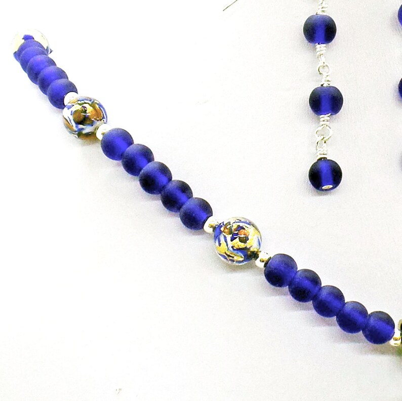 May include: A blue glass bead necklace with silver accents and two matching earrings. The necklace features two larger beads with a white and gold design.