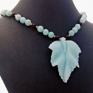 May include: A necklace with a carved green leaf pendant. The necklace is made of green and black beads with copper accents.