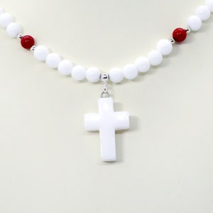 May include: A white beaded necklace with a white cross pendant. The necklace features white and red beads, with silver-colored accents. The cross is a simple, classic design, and the necklace is displayed against a neutral background.