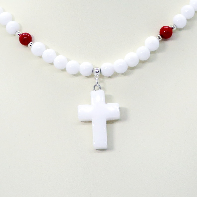 May include: A white beaded necklace with a white cross pendant. The necklace features white and red beads, with silver-colored accents. The cross is a simple, classic design, and the necklace is displayed against a neutral background.