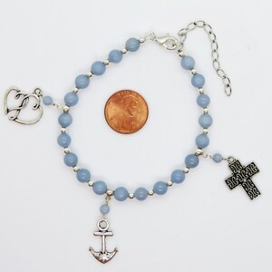 May include: A light blue beaded bracelet with silver-toned charms. Charms include a cross, an anchor, and a double heart. The bracelet is shown on a white background with a penny for size comparison.