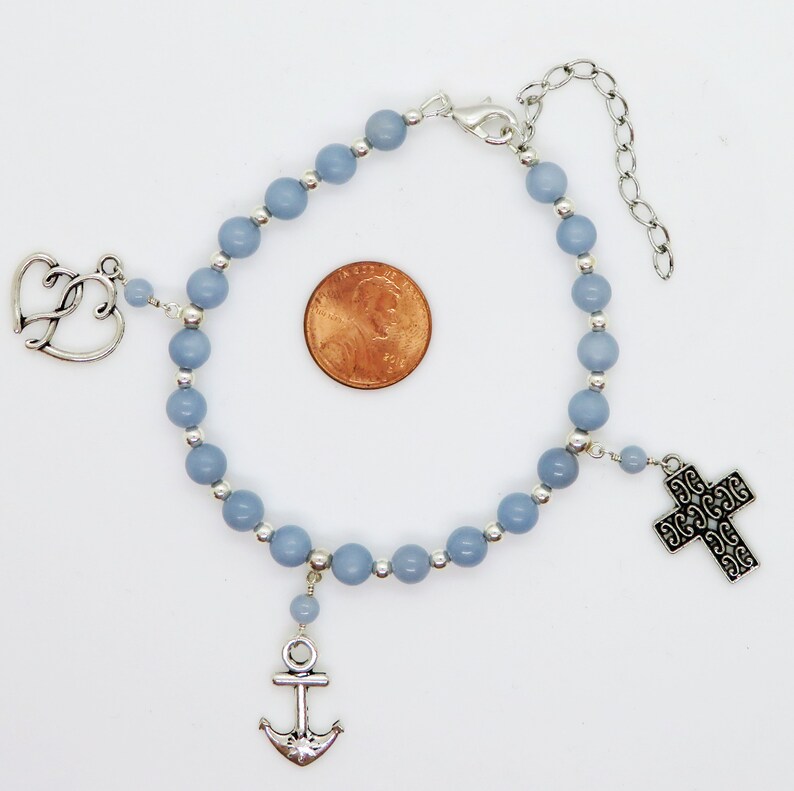 May include: A light blue beaded bracelet with silver-toned charms. Charms include a cross, an anchor, and a double heart. The bracelet is shown on a white background with a penny for size comparison.