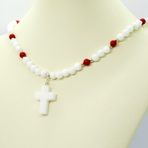 May include: White beaded necklace with a white cross pendant. The necklace features white and red beads, with silver accents. The cross pendant is a solid white color. The necklace is displayed on a mannequin.