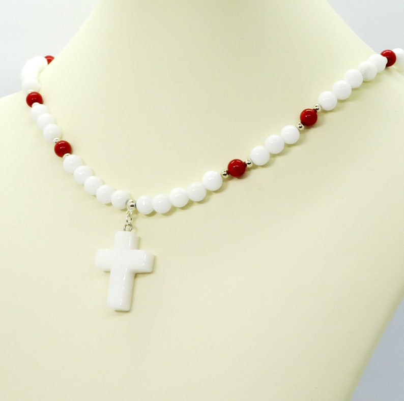 May include: White beaded necklace with a white cross pendant. The necklace features white and red beads, with silver accents. The cross pendant is a solid white color. The necklace is displayed on a mannequin.
