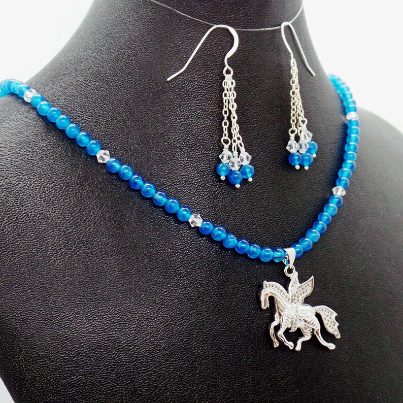 May include: A blue beaded necklace and matching earrings set. The necklace features a silver pendant of a winged horse. The earrings have dangling blue beads and clear crystals. The jewelry is displayed on a black mannequin.