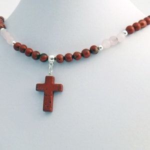 May include: A necklace featuring a red jasper cross pendant. The necklace is strung with red jasper beads, interspersed with pink rose quartz beads and silver accents. The cross is a deep red color.