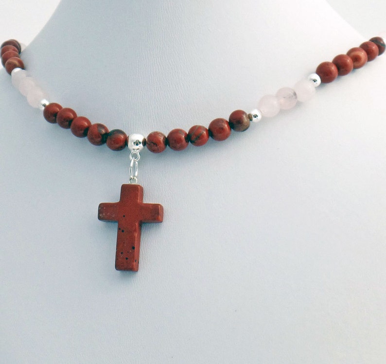 May include: A necklace featuring a red jasper cross pendant. The necklace is strung with red jasper beads, interspersed with pink rose quartz beads and silver accents. The cross is a deep red color.