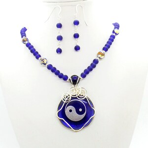 May include: A blue glass necklace with a yin yang pendant. The necklace is made of small blue glass beads and has a silver wire wrapped around the pendant. The pendant is a round blue glass piece with a white yin yang symbol.