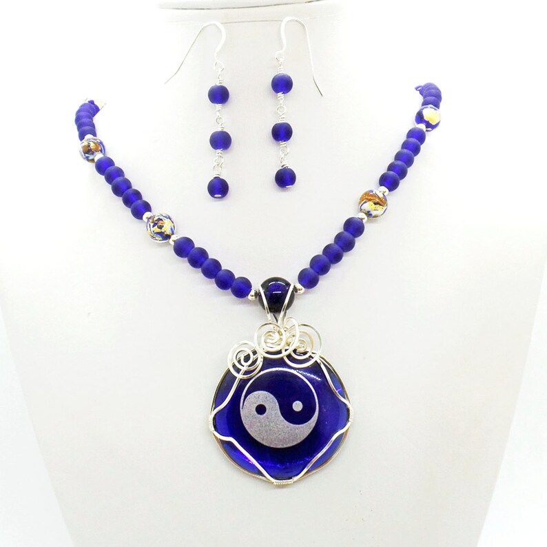 May include: A blue glass necklace with a yin yang pendant. The necklace is made of small blue glass beads and has a silver wire wrapped around the pendant. The pendant is a round blue glass piece with a white yin yang symbol.