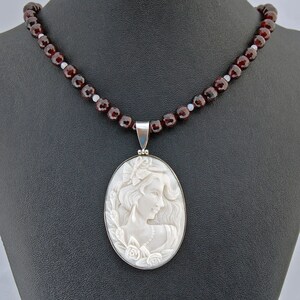 K&ouml;nnte beinhalten: Eine Kamee-Anh&auml;nger-Halskette mit einem geschnitzten Profil einer Frau mit Blumen und flie&szlig;endem Haar. Der Anh&auml;nger ist in einem silbernen Rahmen gefasst und h&auml;ngt an einer Kette aus dunkelroten Granatperlen. Die Halskette wird auf einer schwarzen Oberfl&auml;che pr&auml;sentiert.