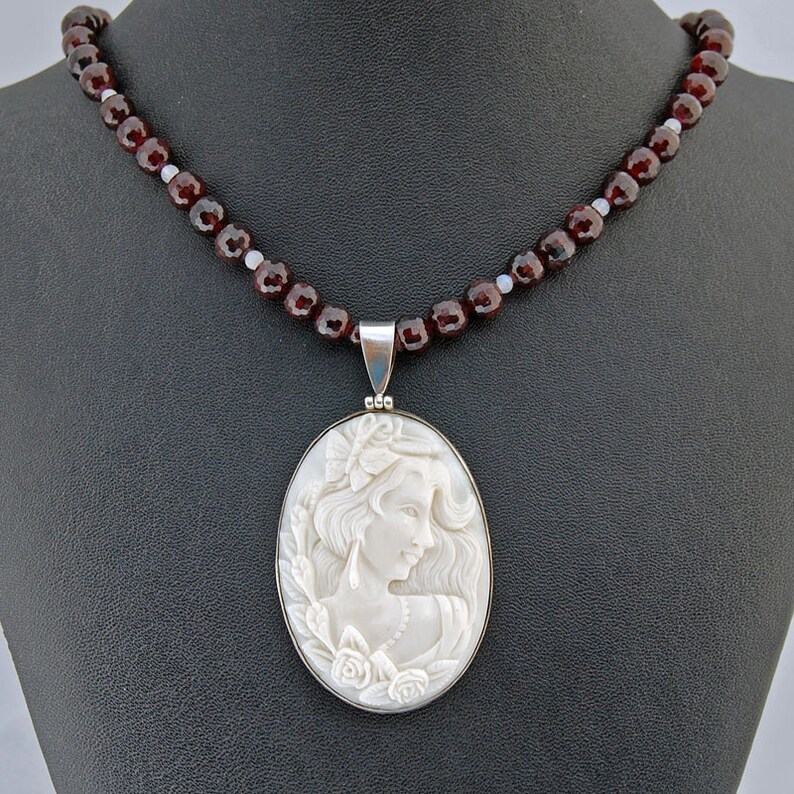 K&ouml;nnte beinhalten: Eine Kamee-Anh&auml;nger-Halskette mit einem geschnitzten Profil einer Frau mit Blumen und flie&szlig;endem Haar. Der Anh&auml;nger ist in einem silbernen Rahmen gefasst und h&auml;ngt an einer Kette aus dunkelroten Granatperlen. Die Halskette wird auf einer schwarzen Oberfl&auml;che pr&auml;sentiert.