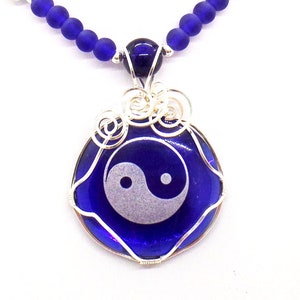 May include: A silver wire wrapped pendant necklace with a blue glass circle featuring a white yin yang symbol. The necklace is made with blue glass beads.